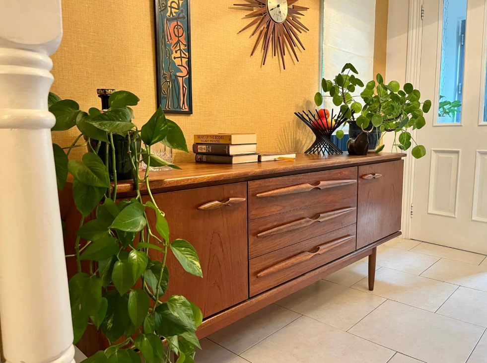 A mid-century modern wooden sideboard with a warm medium brown finish, styled in a home entryway with potted green plants, books and a sunburst wall clock.