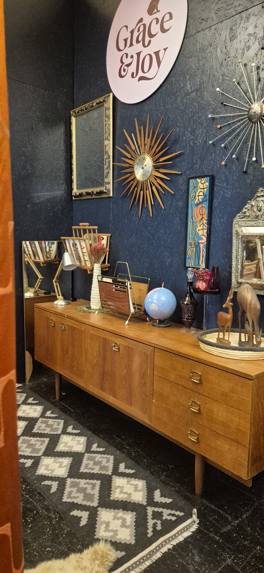 A mid-century modern wooden sideboard cabinet with brass handles, styled in a room with a dark blue textured wall, vintage mirrors, starburst wall decor, and a black-and-white patterned rug.