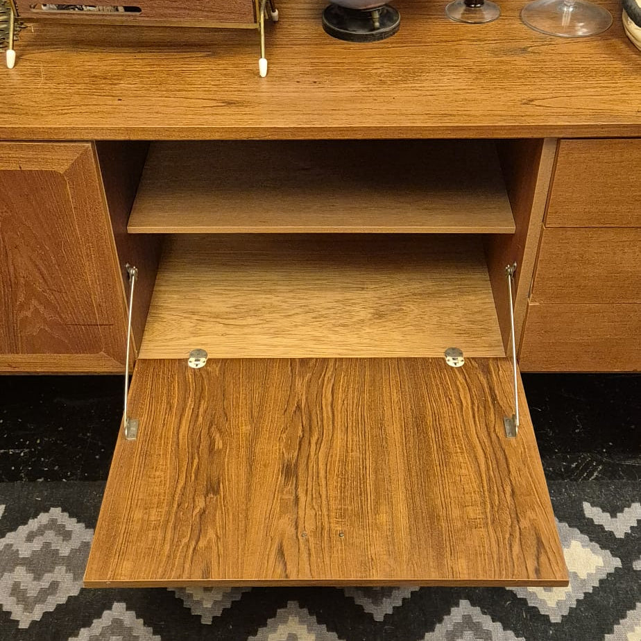 A mid-century modern wooden cabinet with its lower door open, showcasing a warm medium-brown wood grain finish. The cabinet is styled with a globe, a metal magazine rack, and other decor items on top, set against a dark textured wall and a patterned rug.