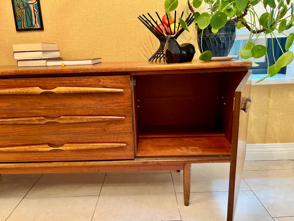 A mid-century modern wooden sideboard with a warm medium brown finish, styled in a home entryway with potted green plants, books and a sunburst wall clock.