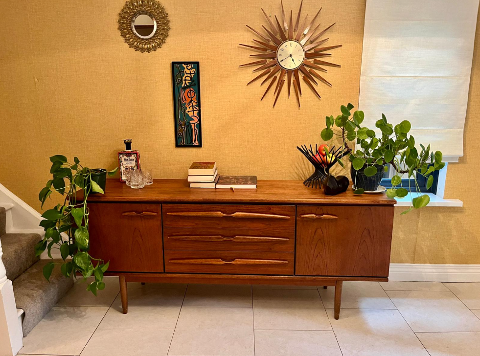 A mid-century modern wooden sideboard with a warm medium brown finish, styled in a home entryway with potted green plants, books and a sunburst wall clock.
