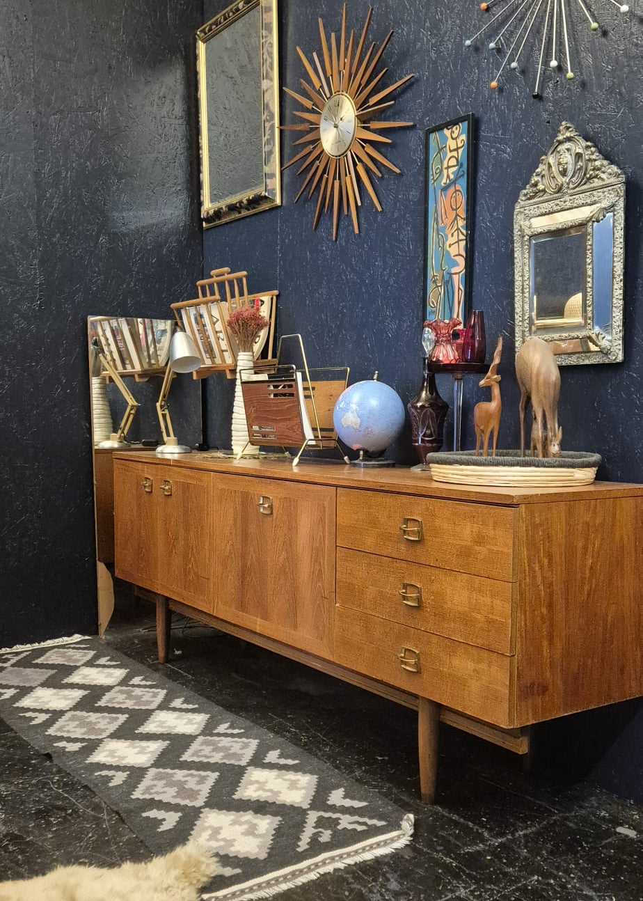 A mid-century modern wooden credenza or sideboard cabinet with six drawers, styled in a dark room with decorative mirrors, starburst wall art, books, and a globe. A patterned rug and a cream-colored faux fur rug are on the floor.