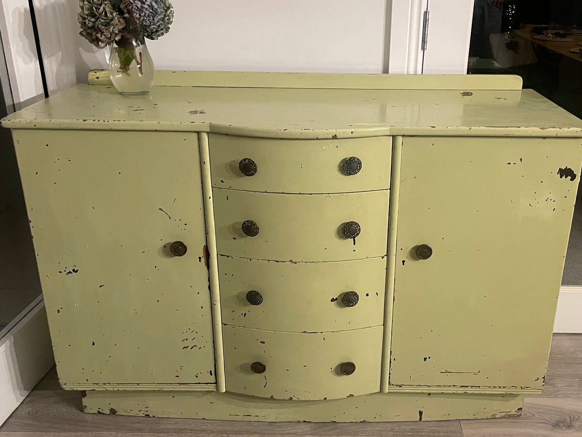 A distressed sage green wooden sideboard or credenza with five drawers in the center and dark round knobs, set against a white wall with glass doors and a wood-look floor.