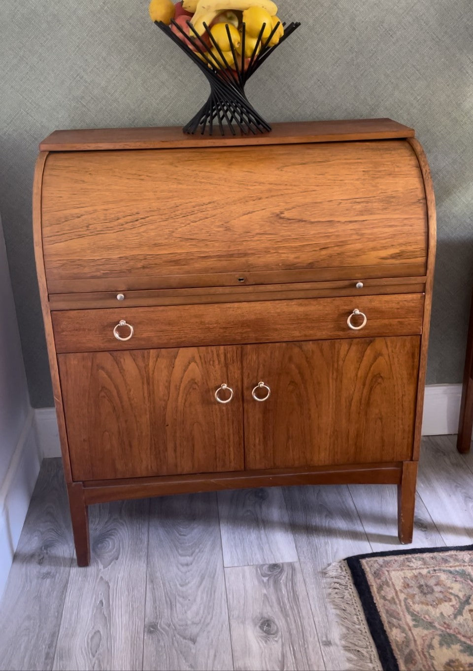 A mid-century modern wooden roll-top desk or secretary cabinet with a warm medium-brown wood grain finish, featuring two drawers on top and two doors below with brass ring pulls, set against a gray wallpapered wall.
