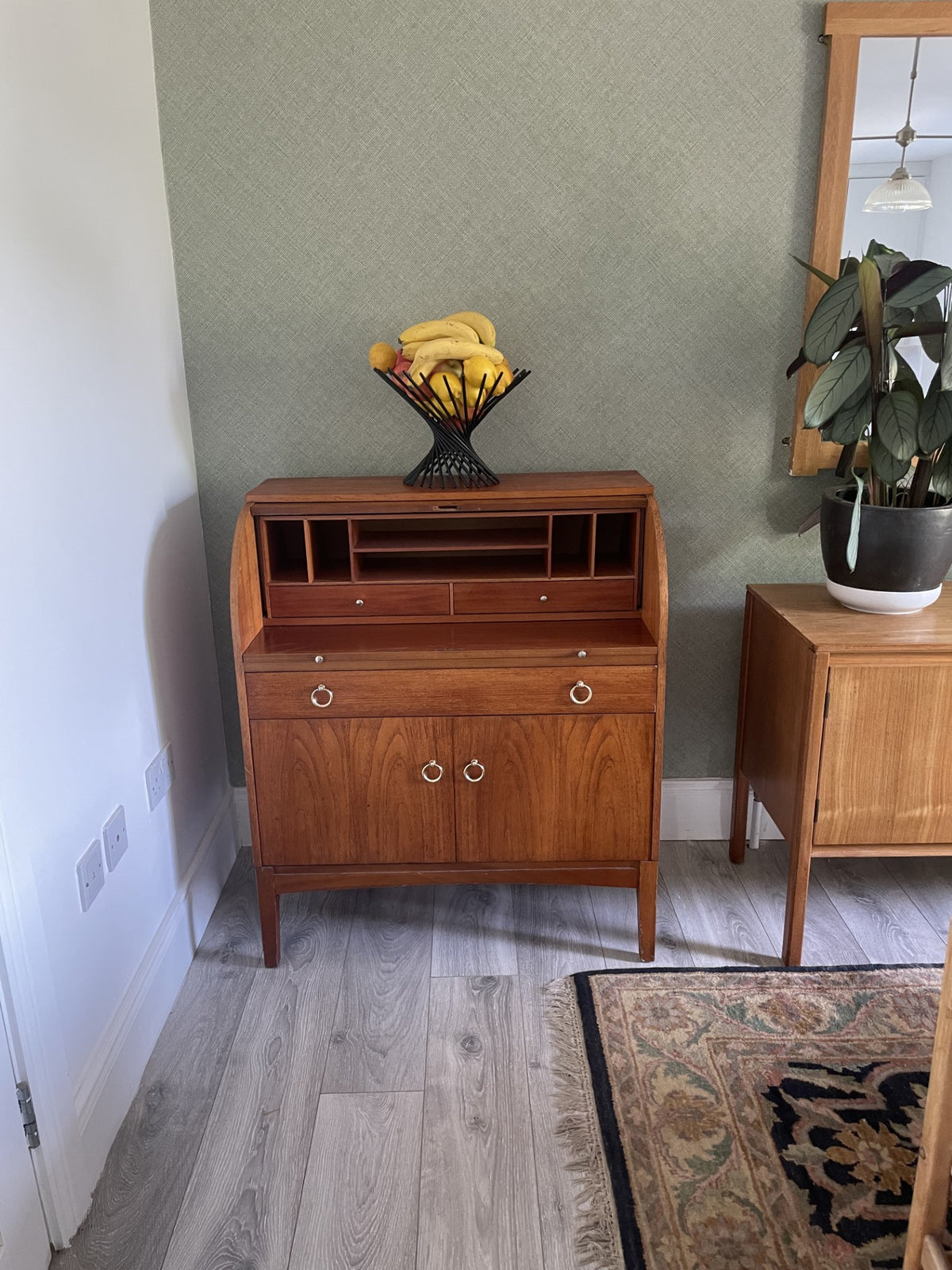 A mid-century modern wooden writing desk or hutch with a warm walnut finish, featuring a roll-top lid, open shelves, two drawers with round metal pulls, and two lower doors with matching pulls. It is styled in a home office setting with a potted plant on an adjacent side table, a green textured wall, and light wood flooring.