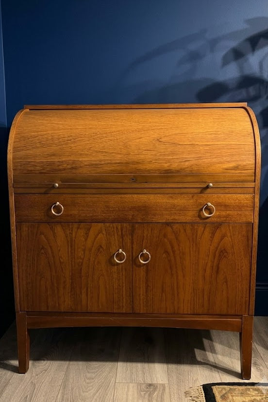 A mid-century remploy wooden roll-top desk cabinet with a warm brown teak wood grain finish, featuring two drawers with brass ring pulls on the upper section and two doors with matching ring pulls on the lower section, set against a dark blue wall and light wood floor.