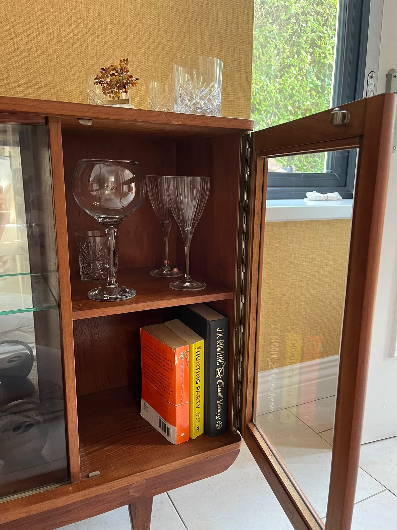 A mid-century modern SF Quinn wooden display cabinet with glass doors, shown in a home interior setting with tiled floors and textured walls by Grace & Joy Furniture
