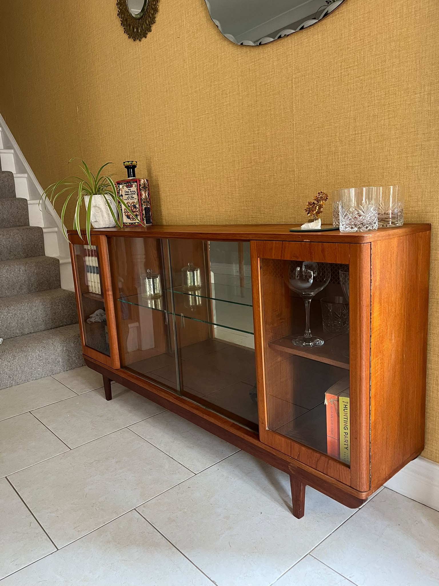 A mid-century modern SF Quinn wooden display cabinet with glass doors, shown in a home interior setting with tiled floors and textured walls by Grace & Joy Furniture