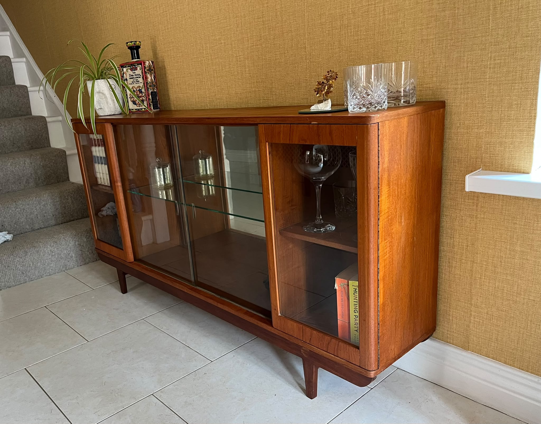 A mid-century modern SF Quinn wooden display cabinet with glass doors, shown in a home interior setting with tiled floors and textured walls by Grace & Joy Furniture