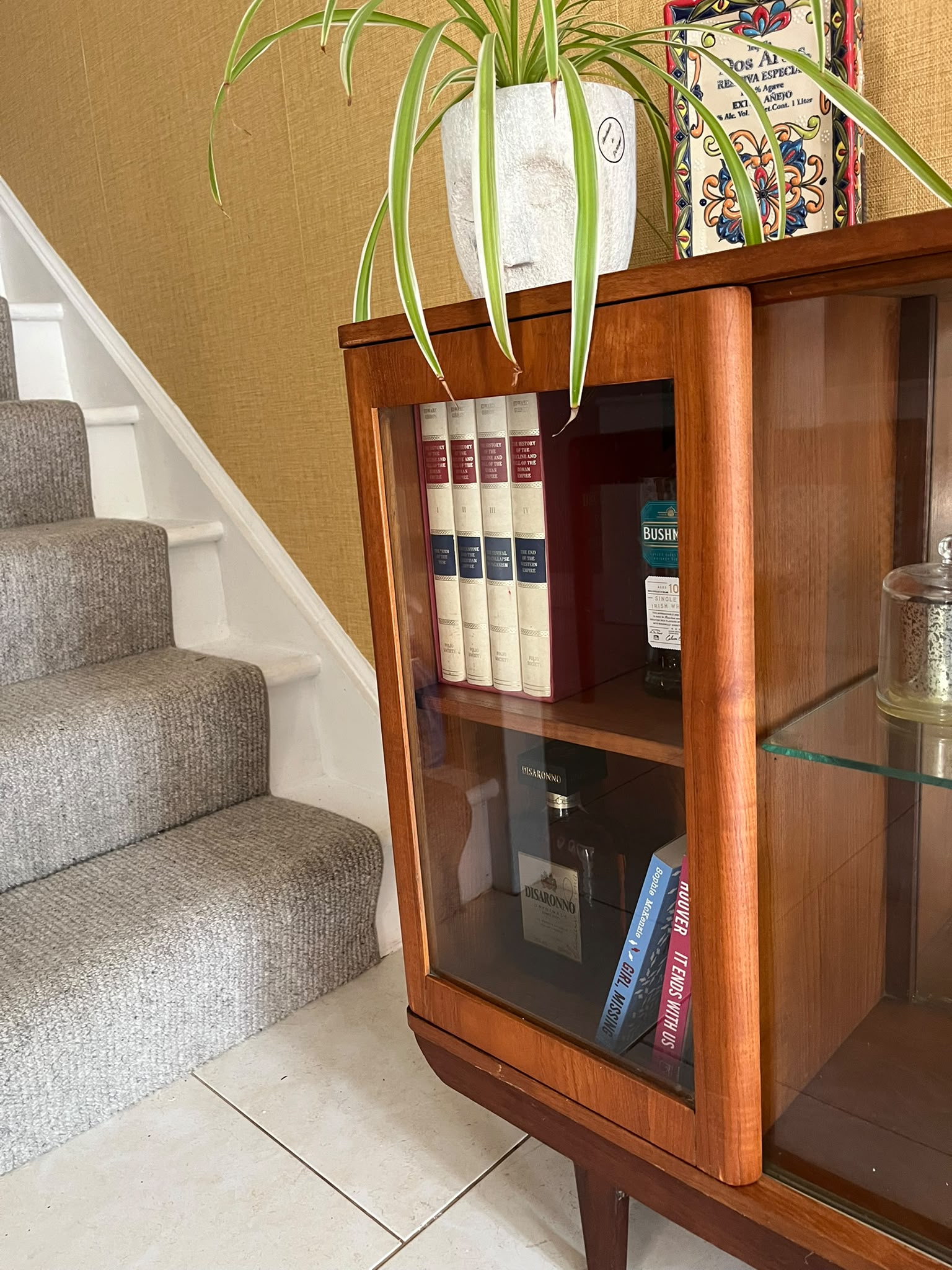 A mid-century modern SF Quinn wooden display cabinet with glass doors, shown in a home interior setting with tiled floors and textured walls by Grace & Joy Furniture