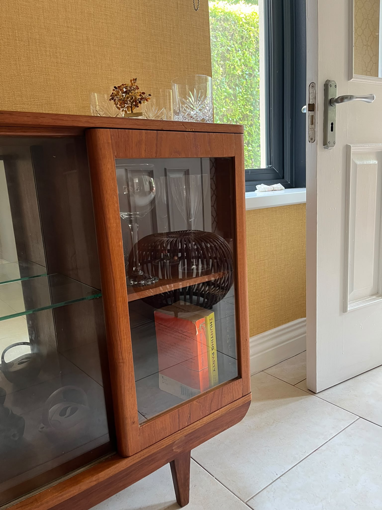 A mid-century modern SF Quinn wooden display cabinet with glass doors, shown in a home interior setting with tiled floors and textured walls by Grace & Joy Furniture