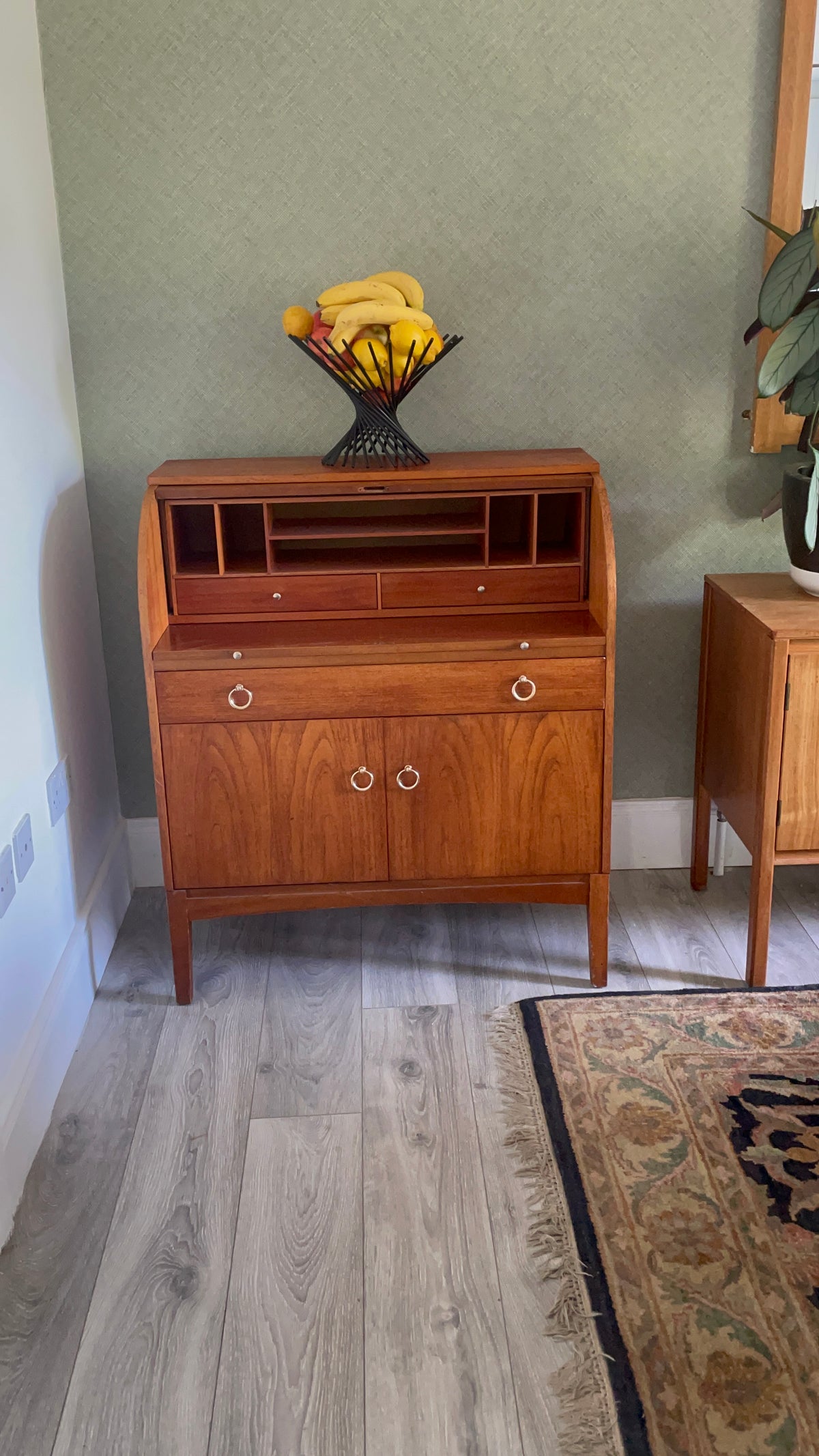 A mid-century modern wooden secretary desk with a warm reddish-brown finish, featuring two drawers on the bottom with round metal pulls, open shelves above, and a roll-top section. It is styled in a room with a textured sage green wall, light wood floors, and a patterned rug.