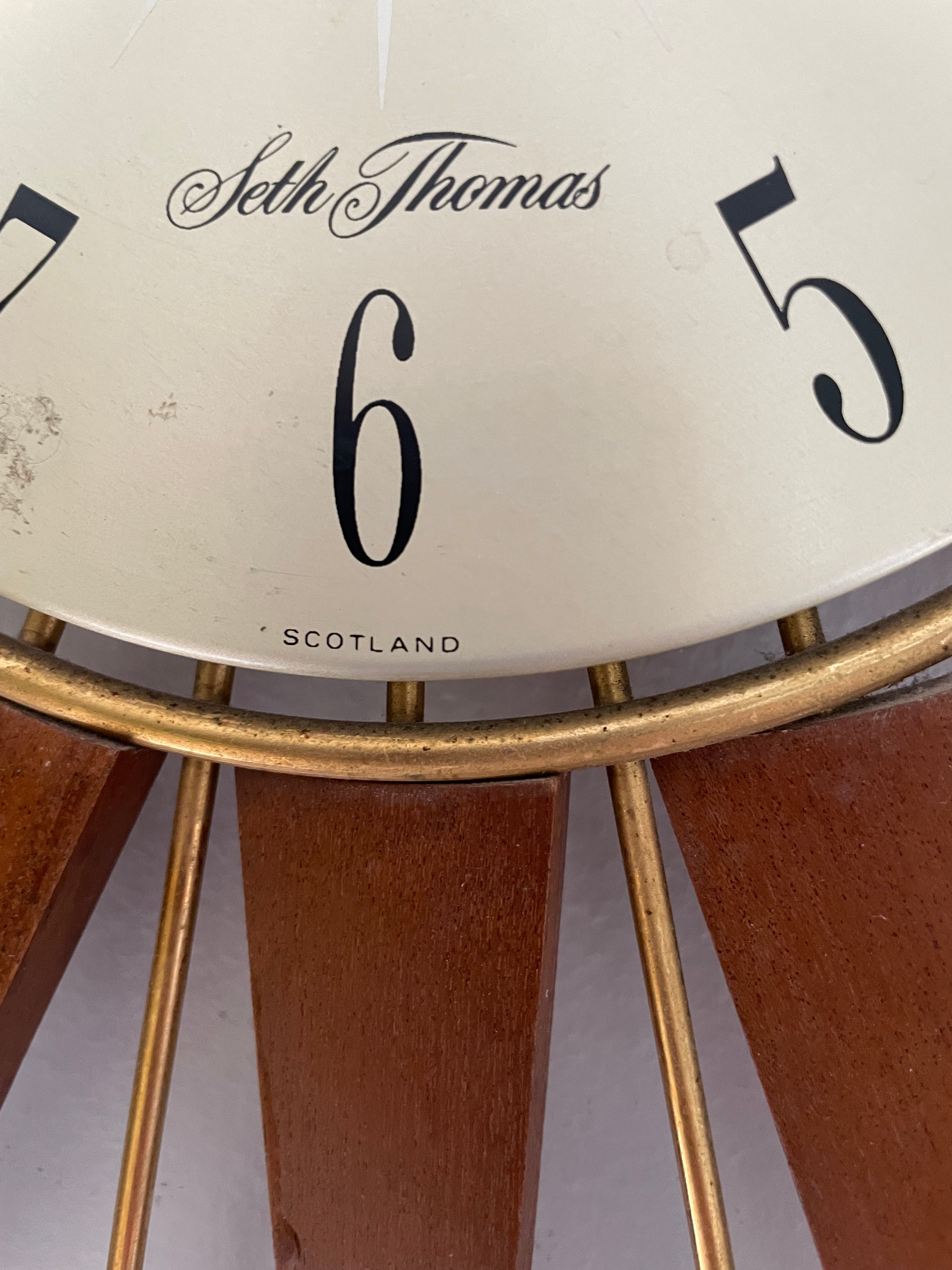 A mid-century modern sunburst wall clock with a circular cream-colored face and numerous radiating wooden clock hands in a warm brown finish, mounted on a plain light gray wall by Grace & Joy