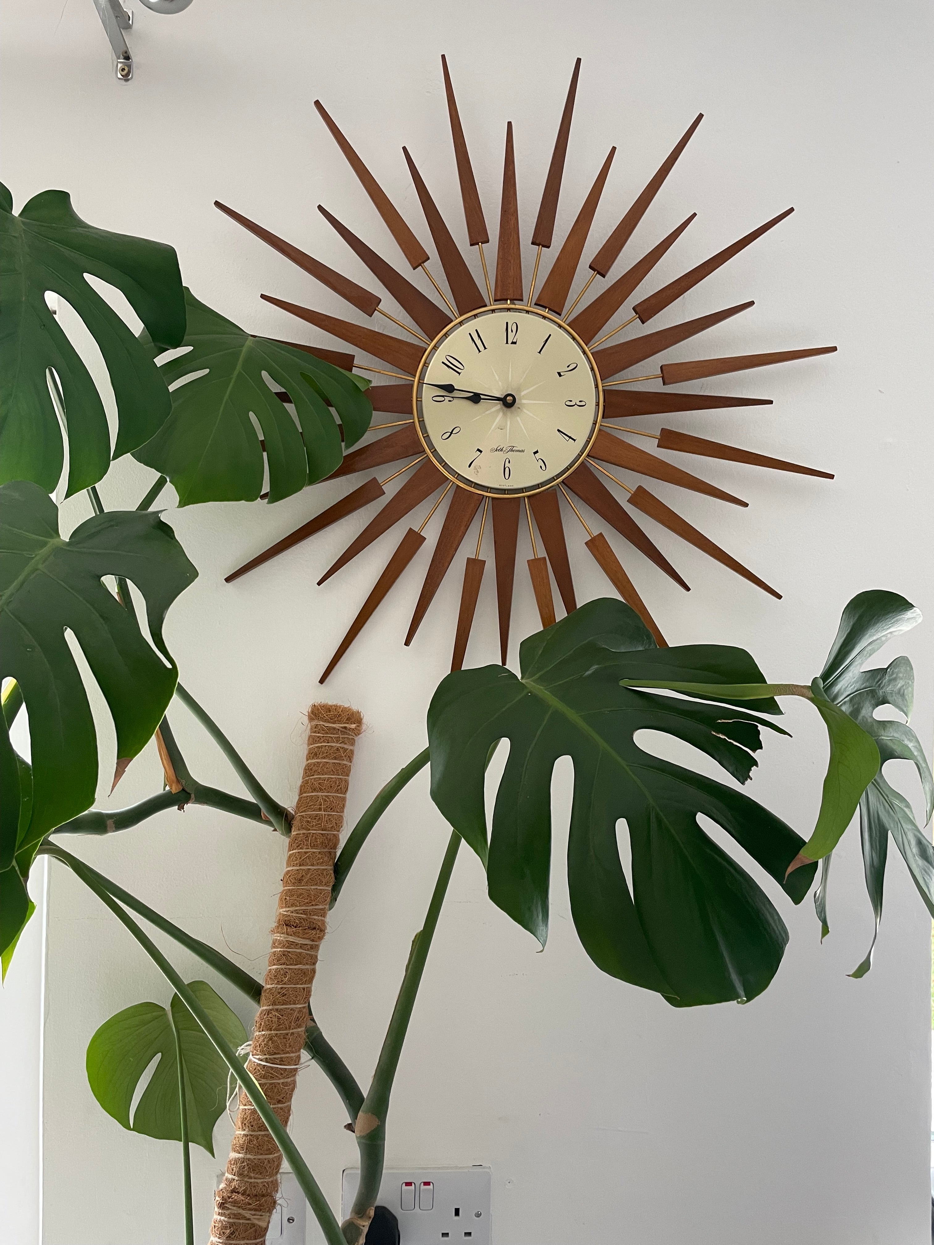 A mid-century modern sunburst wall clock with a circular cream-colored face and numerous radiating wooden clock hands in a warm brown finish, mounted on a plain light gray wall by Grace & Joy