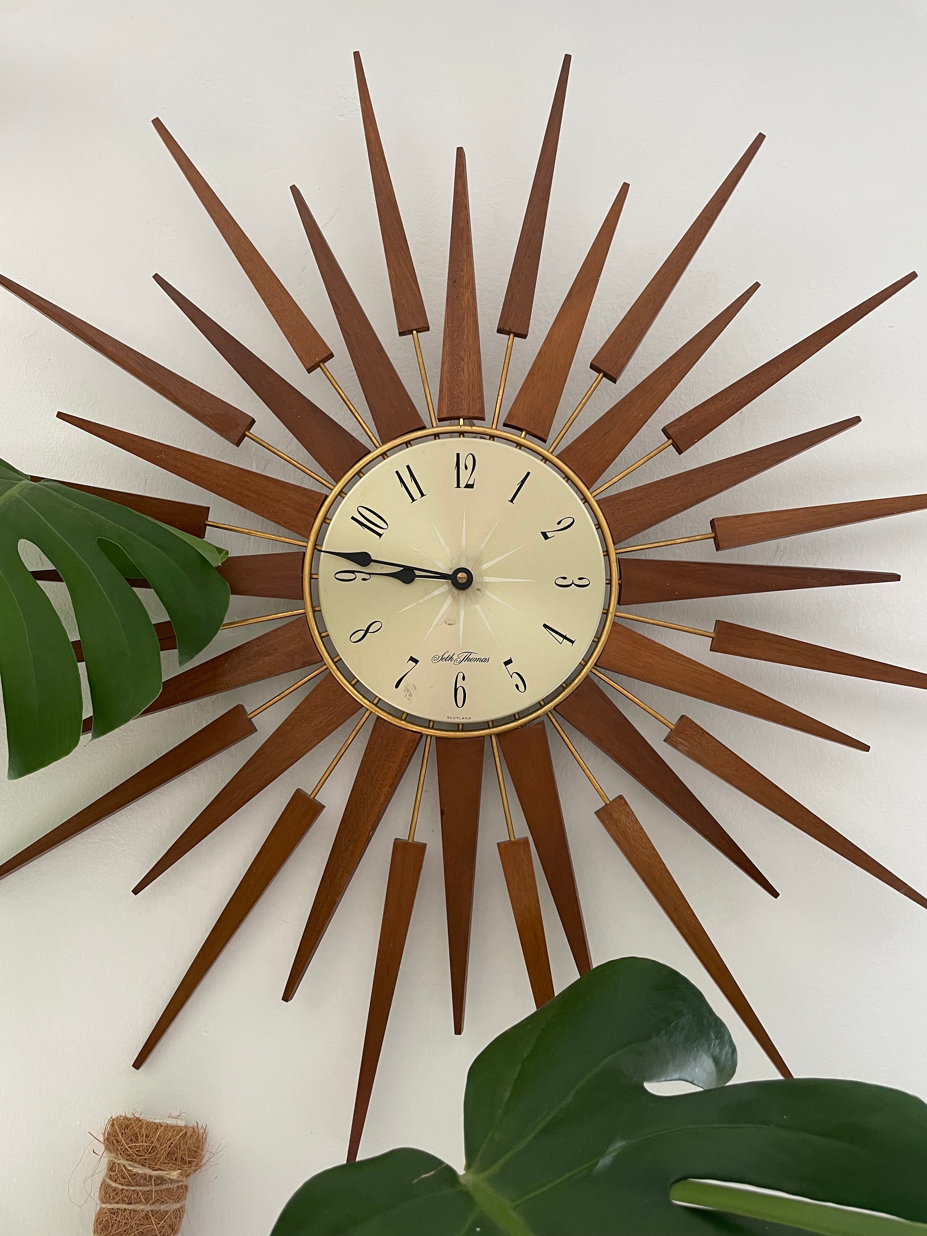 A mid-century modern sunburst wall clock with a circular cream-colored face and numerous radiating wooden clock hands in a warm brown finish, mounted on a plain light gray wall by Grace & Joy