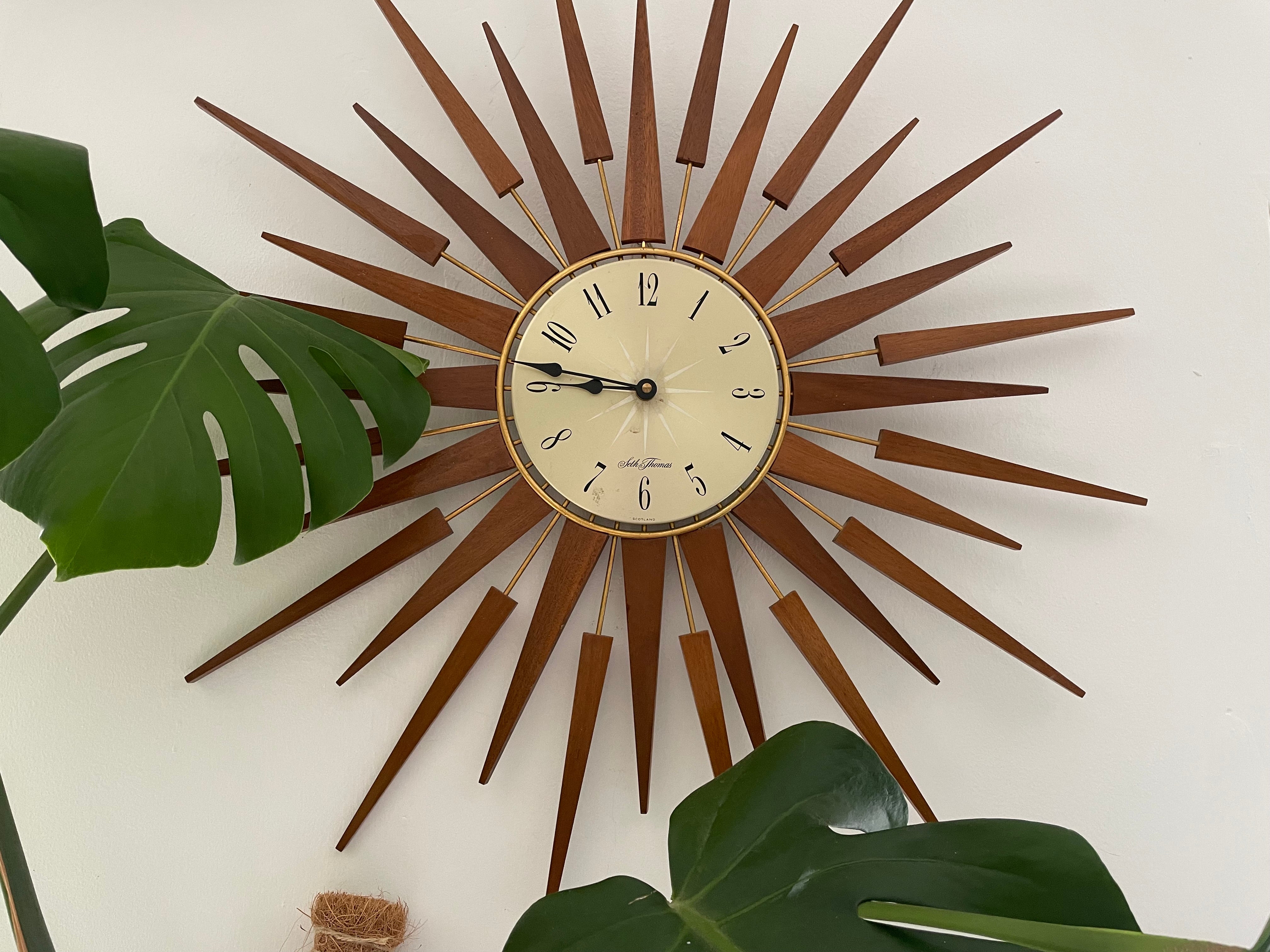 A mid-century modern sunburst wall clock with a circular cream-colored face and numerous radiating wooden clock hands in a warm brown finish, mounted on a plain light gray wall by Grace & Joy
