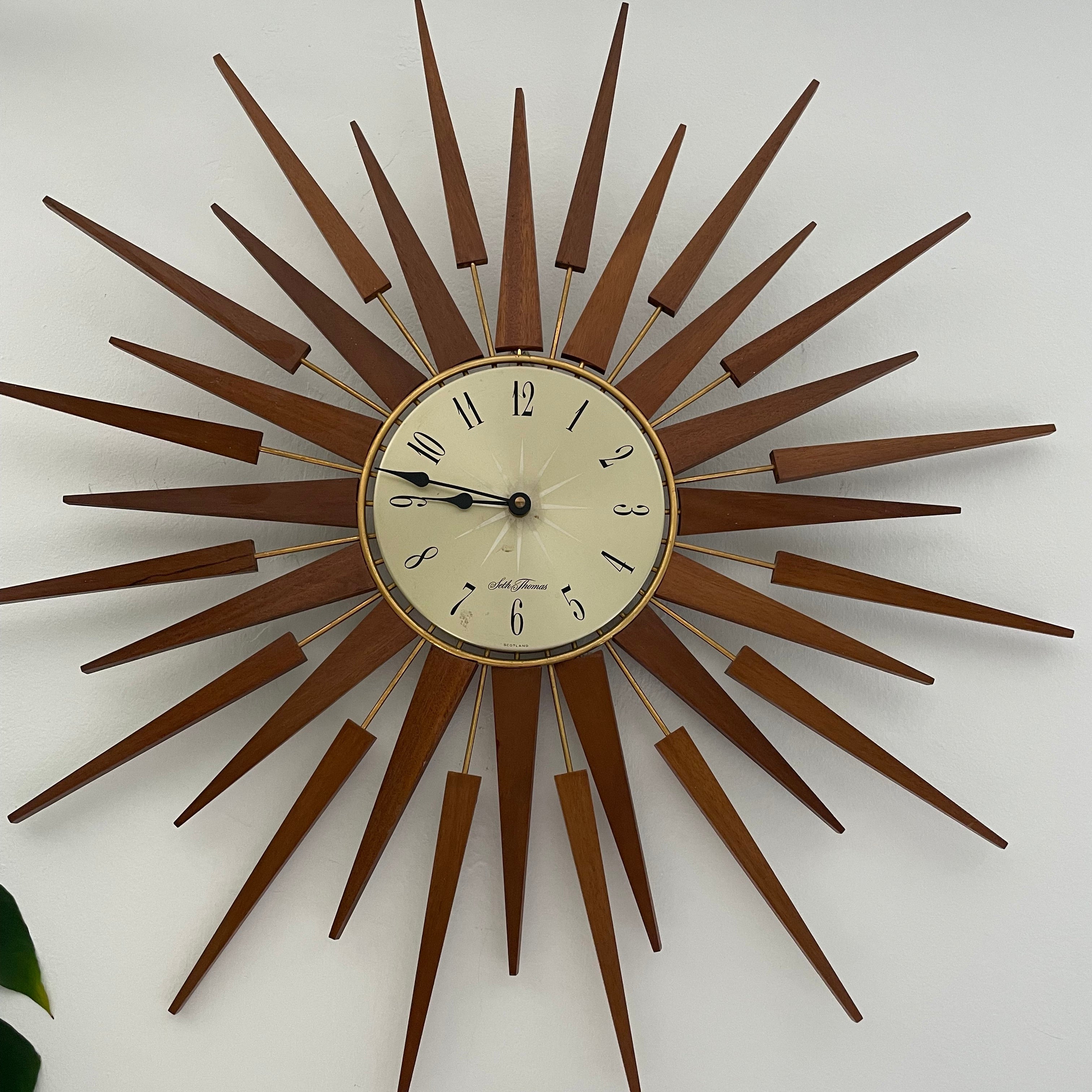A mid-century modern sunburst wall clock with a circular cream-colored face and numerous radiating wooden clock hands in a warm brown finish, mounted on a plain light gray wall by Grace & Joy