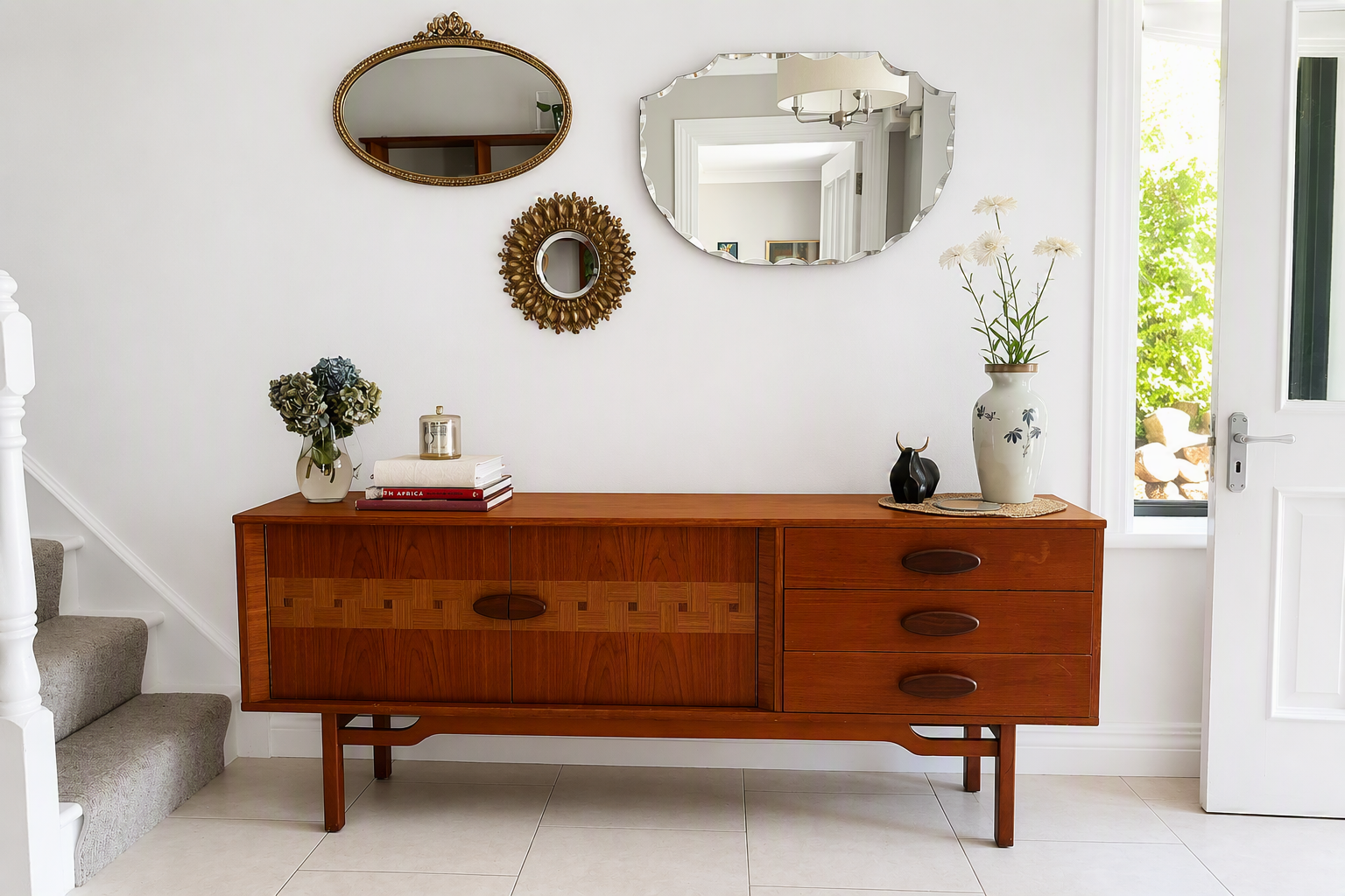 A mid-century modern wooden sideboard or credenza in a warm reddish-brown finish, styled in a bright entryway with decorative mirrors, books, and flowers.