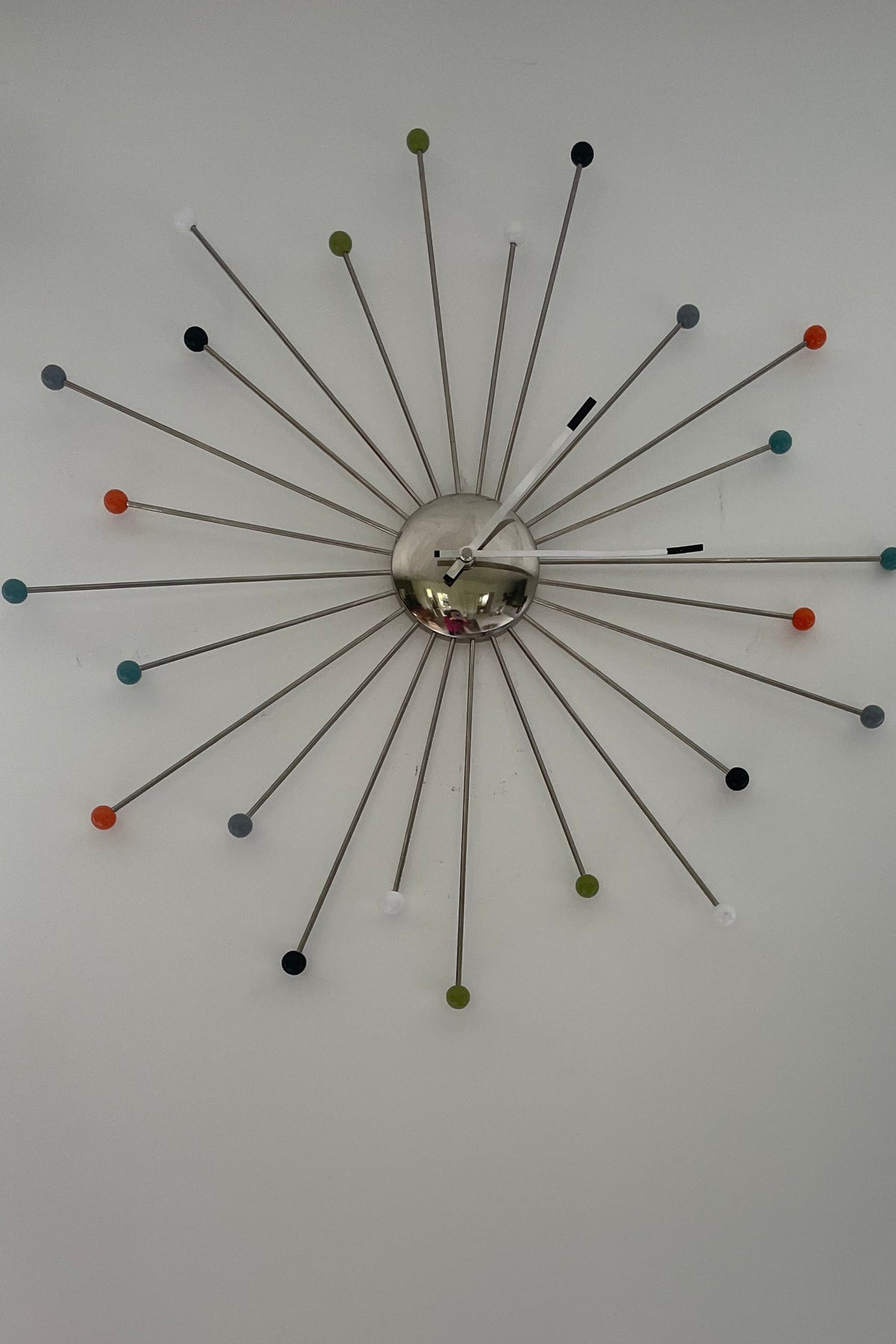 A mid-century modern style starburst wall clock with a polished chrome center and multiple thin metal arms ending in colorful spherical tips, mounted on a plain white wall.
