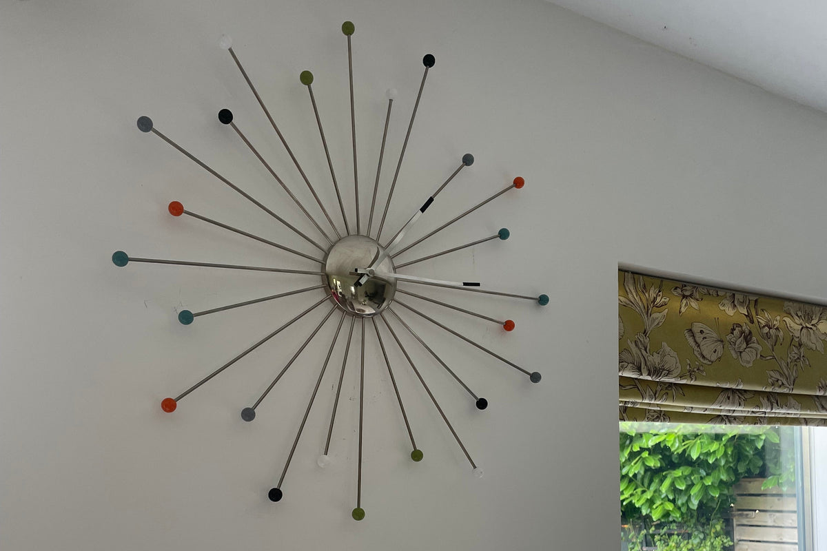 A mid-century style starburst wall clock with a silver metal frame and colorful spherical tips, mounted on a white wall next to a window showing a garden view.
