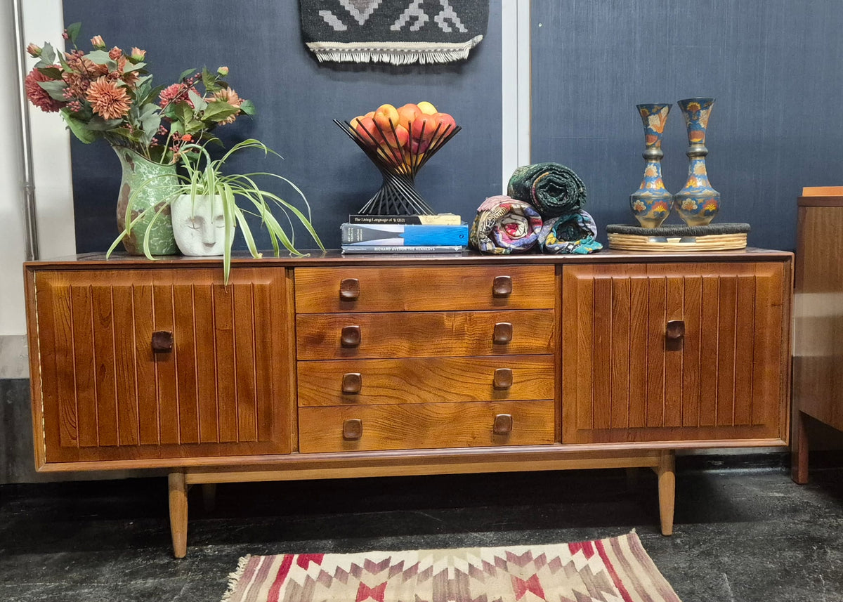 A mid-century stained elm modern wooden sideboard cabinet with three doors and five drawers, styled with home decor items on top against a dark blue wall