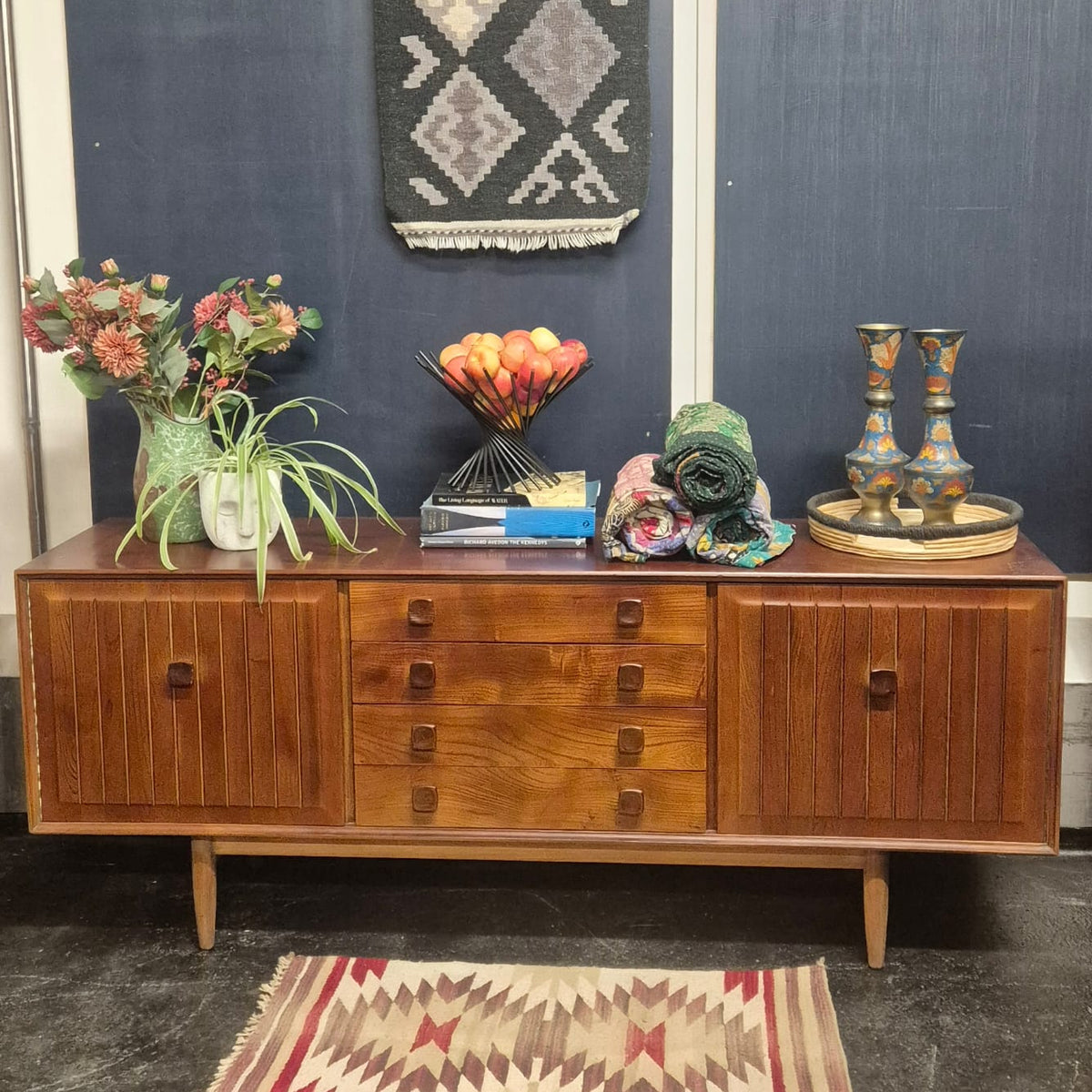 A mid-century stained elm modern wooden sideboard cabinet with three drawers and two cabinet doors, styled with home decor items against a dark blue wall.