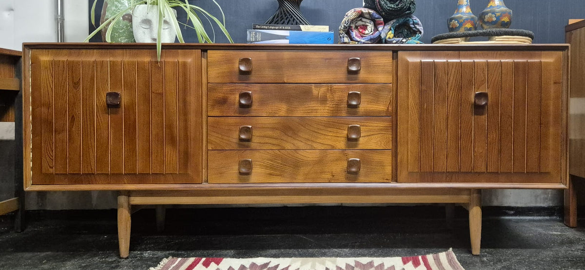 Mid-century modern wooden sideboard cabinet with vertical panel doors and multiple drawers, shown in a room setting with decorative items on top and a patterned rug in front.
