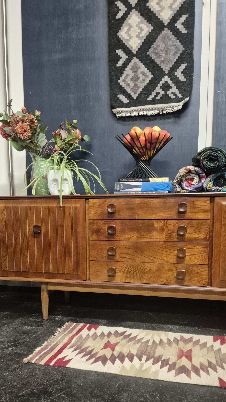 A mid-century modern wooden sideboard cabinet with a warm medium-brown finish, featuring a cabinet door on the left and three drawers on the right with round metal knobs. The cabinet is styled in a room setting with a dark blue wall, a patterned rug, and decorative items on top.