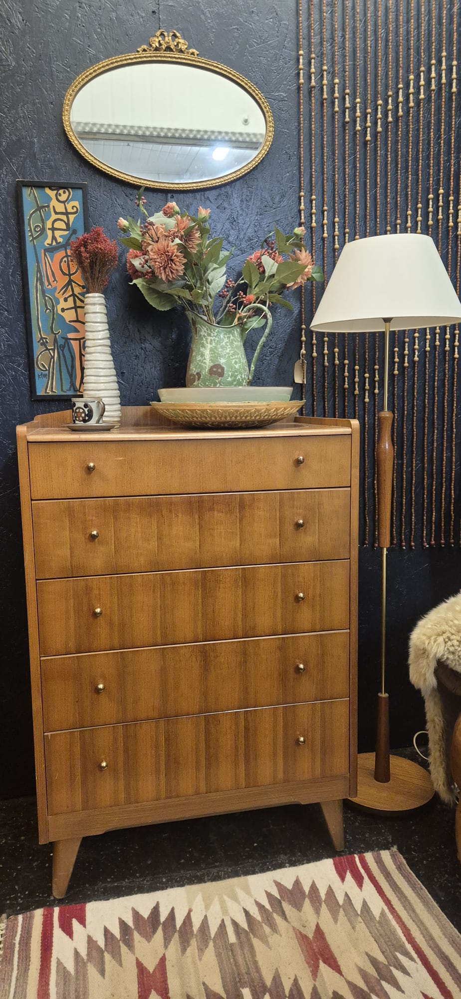 A mid-century modern wooden five-drawer chest of drawers in a warm honey finish, styled in a room with a dark blue textured wall, a gold-framed mirror, a patterned rug, and a floor lamp.