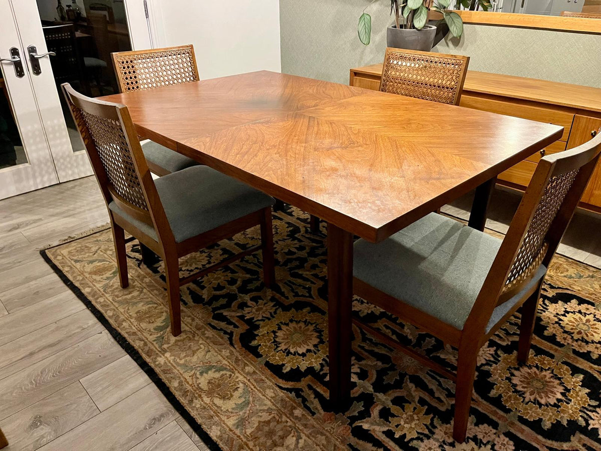 A mid-century modern wooden dining table with four matching woven-back chairs, set on a patterned area rug in a home dining room.