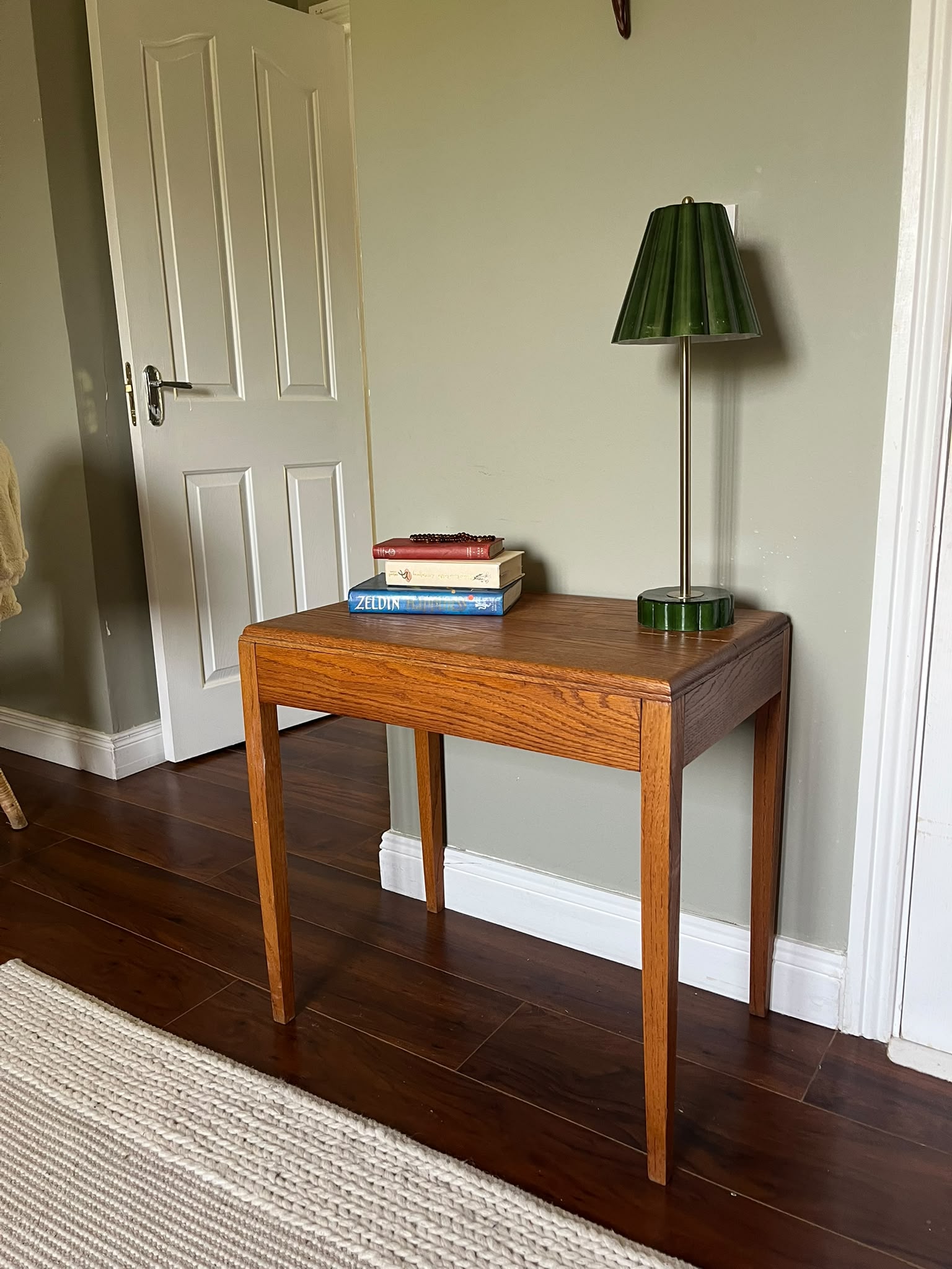 A mid-century modern wooden side table with a warm medium-brown finish, styled with a green pleated table lamp and a stack of books in a room with dark wood floors and a sage green wall by Grace & Joy furniture