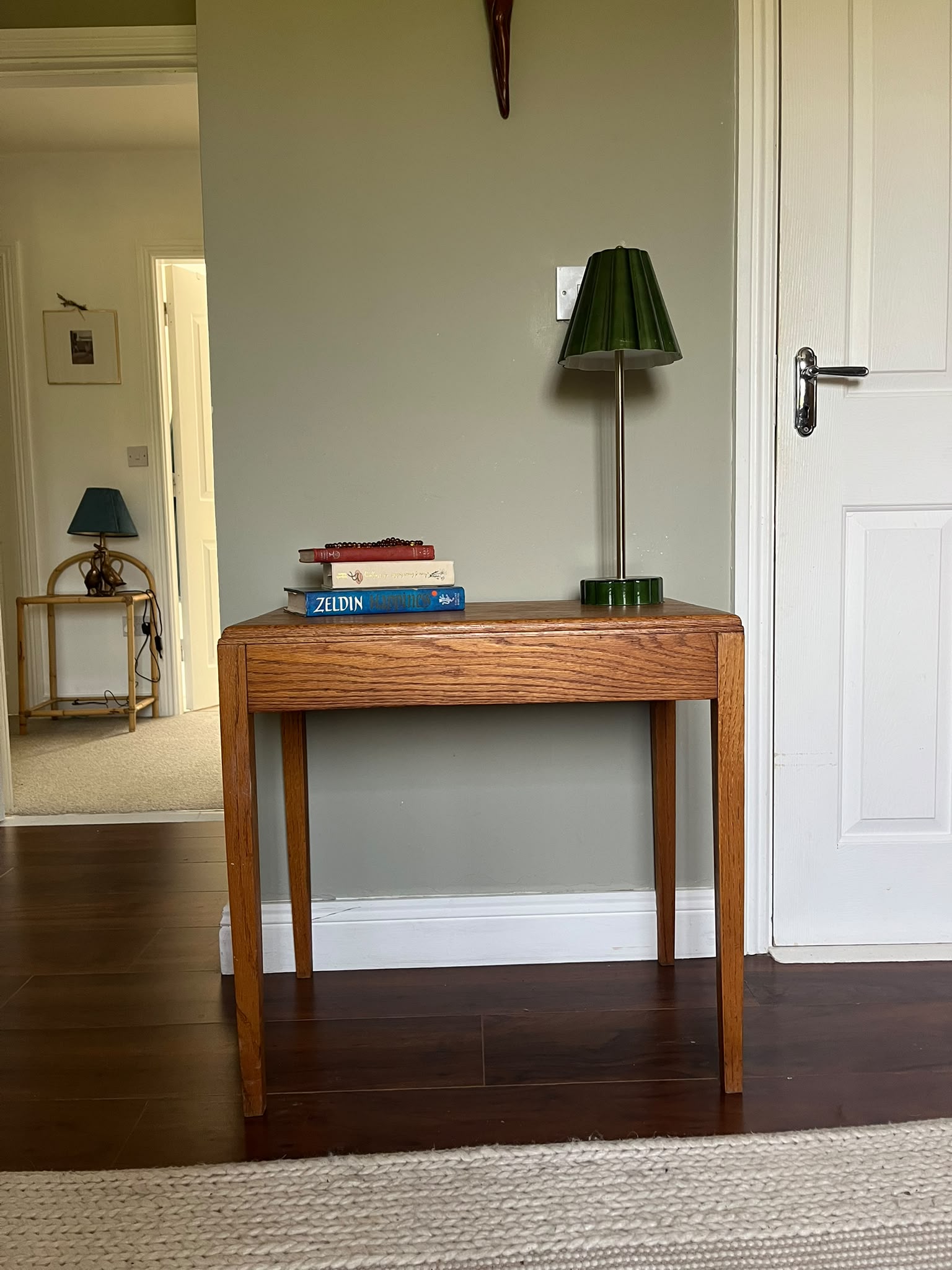 A mid-century modern wooden side table with a warm medium-brown finish, styled with a green pleated table lamp and a stack of books in a room with dark wood floors and a sage green wall by Grace & Joy furniture