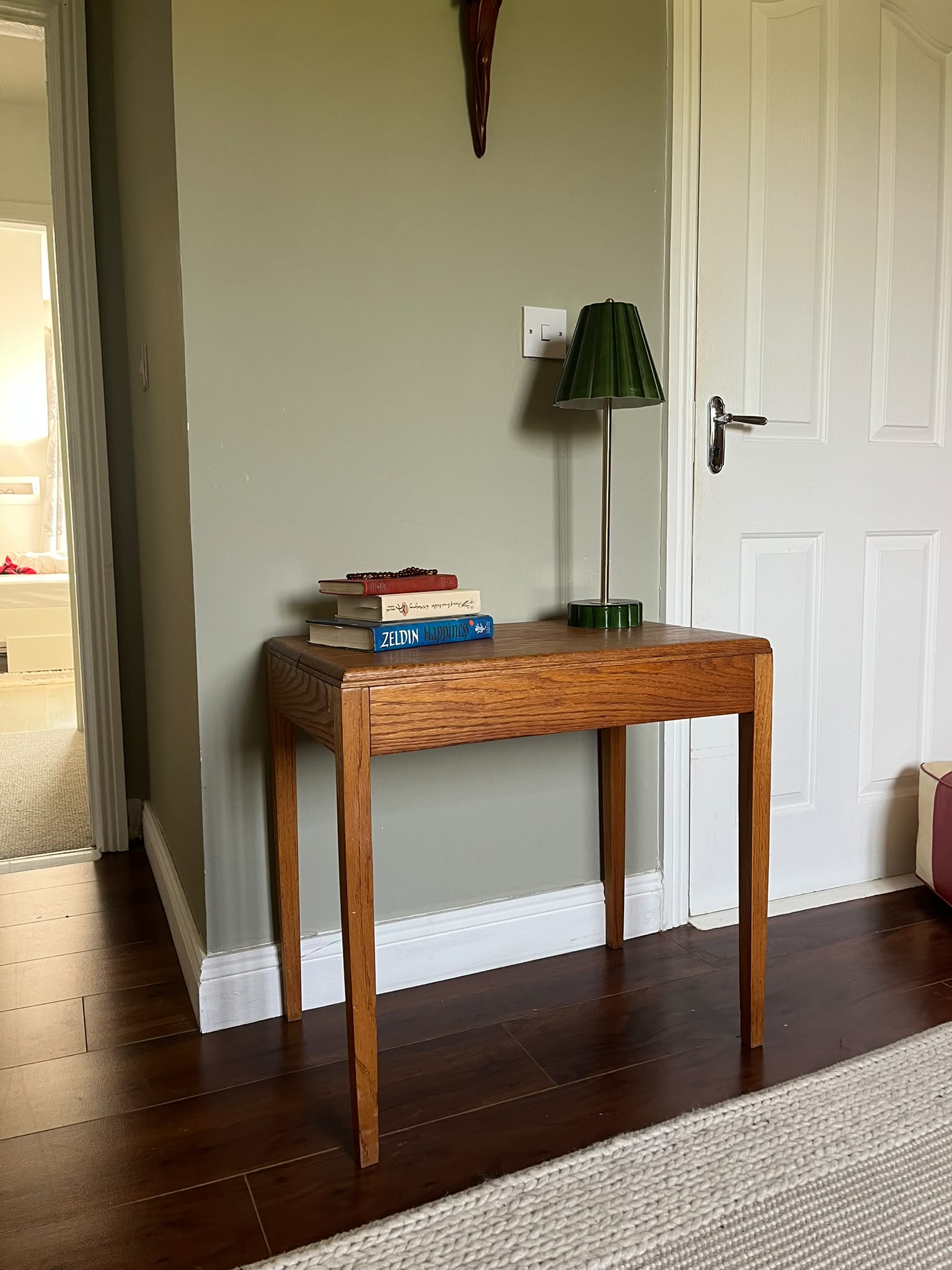A mid-century modern wooden side table with a warm medium-brown finish, styled with a green pleated table lamp and a stack of books in a room with dark wood floors and a sage green wall by Grace & Joy furniture