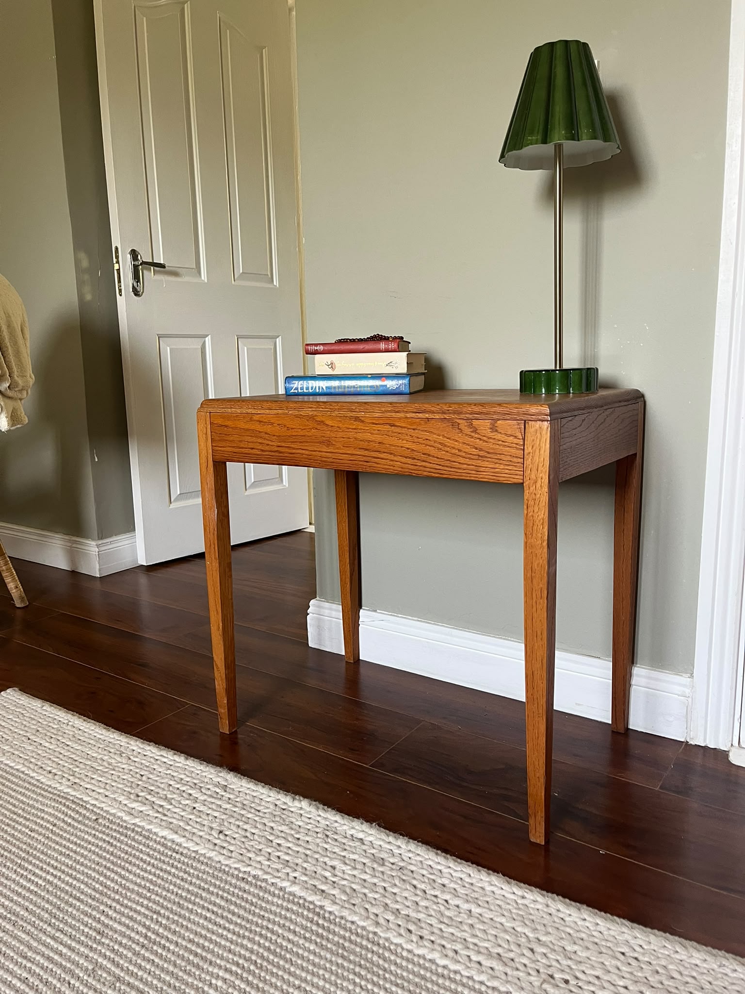 A mid-century modern wooden side table with a warm medium-brown finish, styled with a green pleated table lamp and a stack of books in a room with dark wood floors and a sage green wall by Grace & Joy furniture