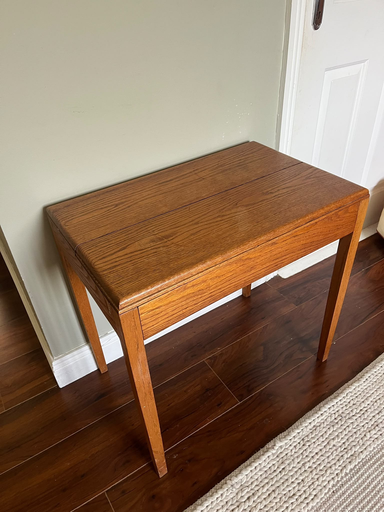 A mid-century modern wooden side table with a warm medium-brown finish, styled with a green pleated table lamp and a stack of books in a room with dark wood floors and a sage green wall by Grace & Joy furniture
